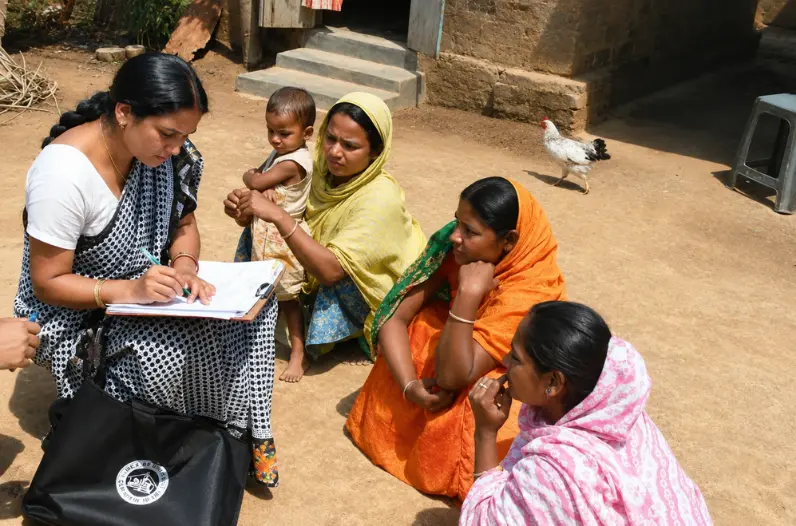 Community worker in a saree writing on a clipboard while conducting a field survey with rural women sitting outdoors.